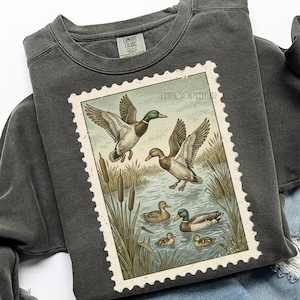 May include: Dark gray sweatshirt featuring a vintage-style stamp illustration of ducks in a pond. The stamp design includes ducks in flight and swimming, with cattails and the text "THE COPTIER". The sweatshirt has a crew neck and long sleeves.