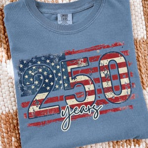 Puede incluir: Camiseta azul con un dise&ntilde;o desgastado de la bandera estadounidense. El gr&aacute;fico incluye el n&uacute;mero "250" y la palabra "years" en cursiva. La camiseta est&aacute; hecha de un material suave y tiene cuello redondo.