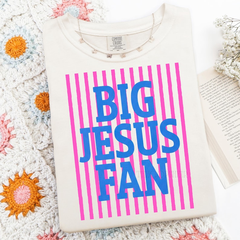 K&ouml;nnte beinhalten: Cremefarbenes T-Shirt mit dem Text "BIG JESUS FAN" in blauen Buchstaben, vor einem Hintergrund aus pinkfarbenen vertikalen Streifen. Eine zarte silberne Halskette mit kleinen Anh&auml;ngern liegt auf dem Shirt. Das Shirt liegt auf einer wei&szlig;en Oberfl&auml;che.