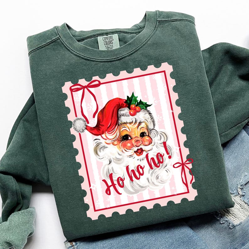 May include: Green crewneck sweatshirt featuring a vintage Santa Claus graphic within a pink postage stamp design. The Santa illustration includes a red hat, white beard, and the words "Ho ho ho!" in red. The sweatshirt has a Comfort Colors tag.