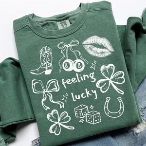 May include: A green sweatshirt with white graphic designs, including a cowboy boot, cherries, lips, shamrocks, dice, a horseshoe, and bows. The words "feeling lucky" are also printed on the shirt.