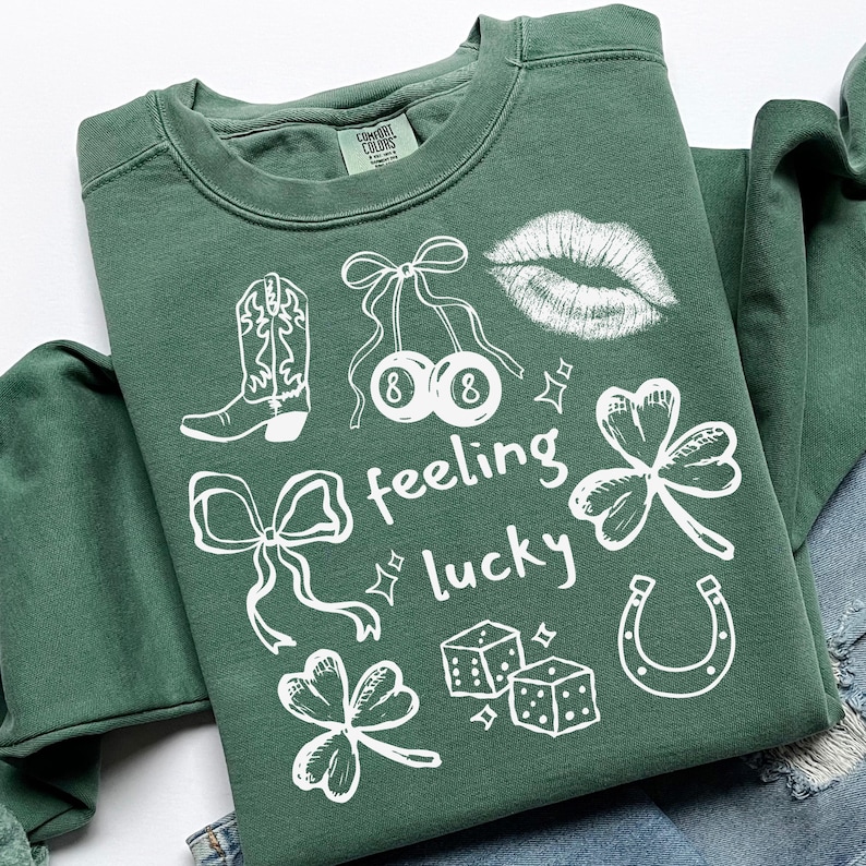 May include: A green sweatshirt with white graphic designs, including a cowboy boot, cherries, lips, shamrocks, dice, a horseshoe, and bows. The words "feeling lucky" are also printed on the shirt.