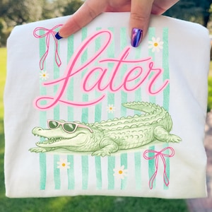 K&ouml;nnte beinhalten: Wei&szlig;es T-Shirt mit einem gr&uuml;nen Alligator mit rosa Sonnenbrille, dem Wort "Later" in rosa Schrift und gr&uuml;n-wei&szlig;en Streifen. Rosa Schleifen und G&auml;nsebl&uuml;mchen-Akzente runden das Design ab.