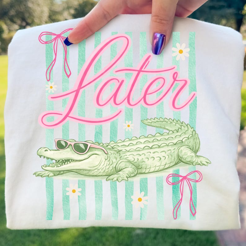 K&ouml;nnte beinhalten: Wei&szlig;es T-Shirt mit einem gr&uuml;nen Alligator mit rosa Sonnenbrille, dem Wort "Later" in rosa Schrift und gr&uuml;n-wei&szlig;en Streifen. Rosa Schleifen und G&auml;nsebl&uuml;mchen-Akzente runden das Design ab.
