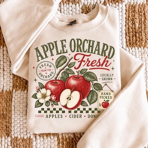 May include: A cream-colored crewneck sweatshirt featuring a graphic with red apples, green leaves, and white blossoms. The text reads APPLE ORCHARD Fresh, FRESH FROM THE ORCHARD, LOCALLY GROWN, HAND PICKED, and APPLES CIDER DONUTS in red and green.