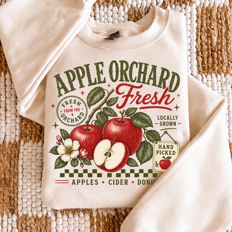 May include: A cream-colored crewneck sweatshirt featuring a graphic with red apples, green leaves, and white blossoms. The text reads APPLE ORCHARD Fresh, FRESH FROM THE ORCHARD, LOCALLY GROWN, HAND PICKED, and APPLES CIDER DONUTS in red and green.