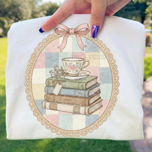 May include: White t-shirt with a vintage design. The design features a stack of books, a teacup, and a bow. The design is framed by a decorative oval border. The text "THECOPYHEIR" is visible.