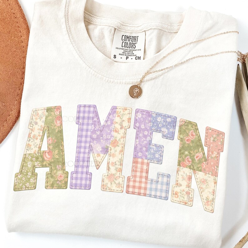 May include: Cream-colored t-shirt with the word "AMEN" in patchwork letters. The letters feature floral and gingham patterns in shades of green, purple, and pink. A gold necklace with a pendant rests on the shirt.