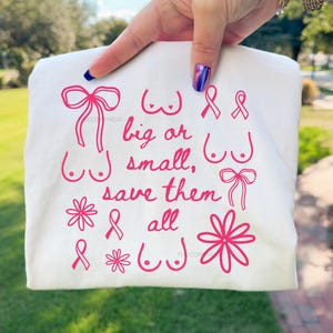 May include: A white t-shirt with pink graphics and text. The design includes illustrations of bows, flowers, and breasts, along with the words "big or small, save them all."