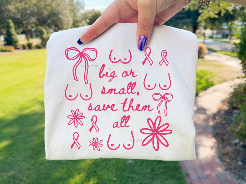 May include: A white t-shirt with pink graphics and text. The design includes illustrations of bows, flowers, and breasts, along with the words "big or small, save them all."