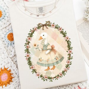 May include: Cream-colored Comfort Colors sweatshirt featuring a whimsical illustration of a goose in a dress holding a flower, framed by a floral wreath. A delicate butterfly necklace rests on the shirt. The design includes the text "DECOPYHEIR".