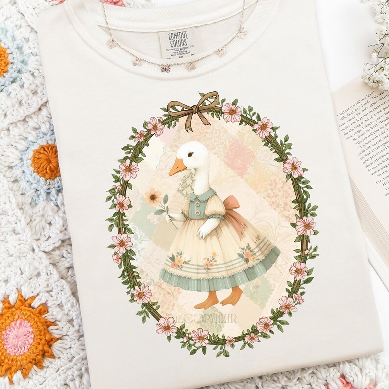 May include: Cream-colored Comfort Colors sweatshirt featuring a whimsical illustration of a goose in a dress holding a flower, framed by a floral wreath. A delicate butterfly necklace rests on the shirt. The design includes the text "DECOPYHEIR".