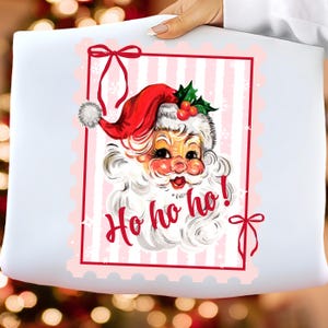 May include: A white pillow with a vintage Santa Claus graphic. The Santa has a red hat, white beard, and rosy cheeks. The words "Ho ho ho!" are written in red script. The design is framed with a red border and pink stripes.