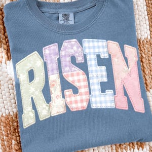 May include: A blue Comfort Colors t-shirt with the word "RISEN" in large, colorful, patchwork-style letters. The letters are in pastel colors and floral patterns. The shirt is on a woven surface.