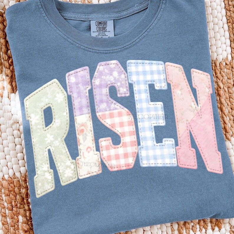 May include: A blue Comfort Colors t-shirt with the word "RISEN" in large, colorful, patchwork-style letters. The letters are in pastel colors and floral patterns. The shirt is on a woven surface.