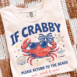 May include: Cream-colored t-shirt with a graphic of a red crab wearing sunglasses and a bow. The text "IF CRABBY PLEASE RETURN TO THE BEACH" is in blue. The shirt also has the words "sun, sand & sass".