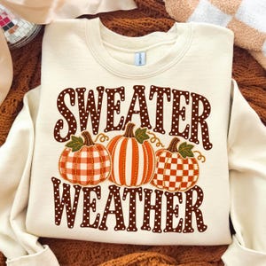 May include: A cream-coloured crewneck sweatshirt featuring the text "SWEATER WEATHER" in brown with white polka dots. Below the text are three pumpkins: one with a red and white gingham pattern, one with orange stripes, and one with a brown checkerboard pattern.