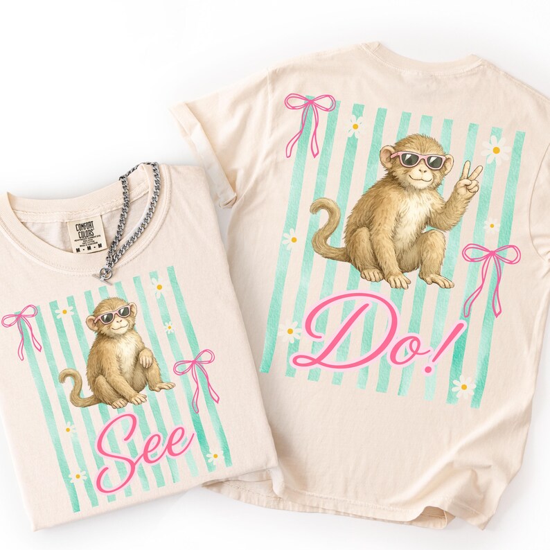 May include: Two cream-colored t-shirts with a monkey graphic wearing sunglasses. The shirts have a light blue striped background with pink bows and the words "See" and "Do!" in pink script. A silver chain necklace is on one shirt.