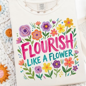 May include: Cream-colored t-shirt with the words "FLOURISH LIKE A FLOWER" in bright pink and teal, surrounded by colorful painted flowers. A delicate butterfly necklace rests on the shirt. The shirt is from Comfort Colors.