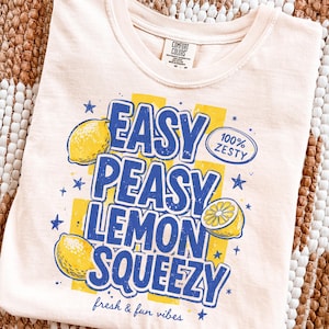 Może przedstawiać: Kremowa koszulka z zabawną grafiką z motywem cytryny. Projekt zawiera tekst "Easy Peasy Lemon Squeezy" w kolorze niebieskim, z żółtymi ilustracjami i akcentami cytryny. Koszulka zawiera również frazę "fresh & fun vibes".