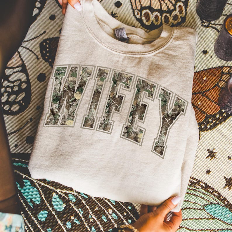 May include: A beige sweatshirt with the word "WIFEY" in large, block letters across the chest. The letters are filled with a camouflage pattern and outlined in a light gray. The sweatshirt is folded and held by hands.