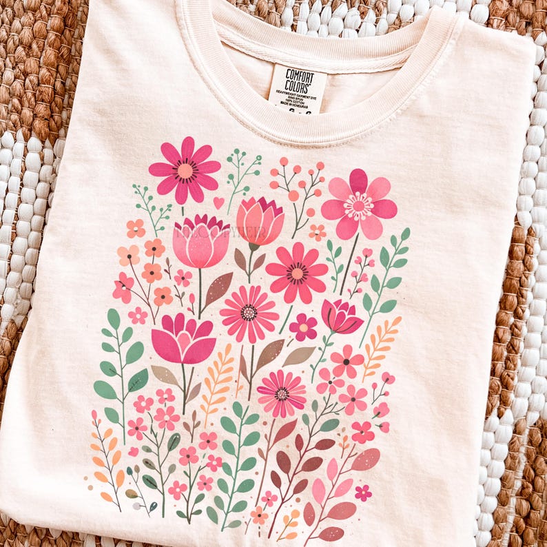May include: Cream-colored t-shirt featuring a vibrant floral design. The design includes pink flowers, green leaves, and brown stems. The shirt has a crew neck and short sleeves. The tag reads "Comfort Colors".