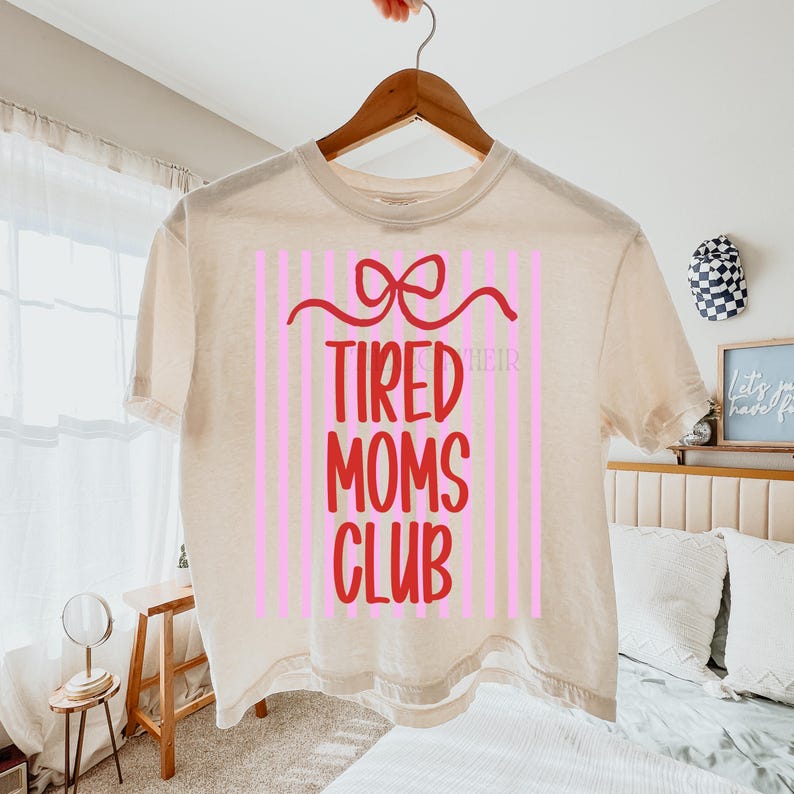 K&ouml;nnte beinhalten: Ein cremefarbenes T-Shirt mit dem Text "TIRED MOMS CLUB" in Rot, vor einem rosa gestreiften Hintergrund. Ein rotes Schleifendesign befindet sich &uuml;ber dem Text. Das Shirt h&auml;ngt an einem Holzb&uuml;gel.