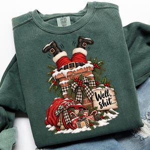 May include: A dark green crewneck sweatshirt featuring a graphic of Santa's legs sticking out of a brick chimney. The design includes a red sack, wrapped gifts, pine branches, and a wooden sign with the text "Well, shit..." in black script.
