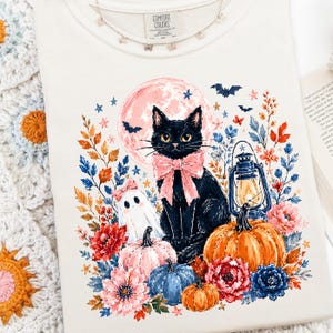 May include: A cream-colored t-shirt featuring a graphic of a black cat with a pink bow, a small ghost, a lantern, pumpkins in orange, pink, and blue, and colorful flowers. The shirt has a Comfort Colors label at the neck and a crew neckline.