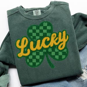 K&ouml;nnte beinhalten: Gr&uuml;nes Sweatshirt mit einem gro&szlig;en, gestickten Kleeblatt-Design. Das Kleeblatt ist gr&uuml;n mit einem Karomuster, und das Wort "Lucky" ist gelb gestickt. Das Sweatshirt ist von der Marke Comfort Colors.
