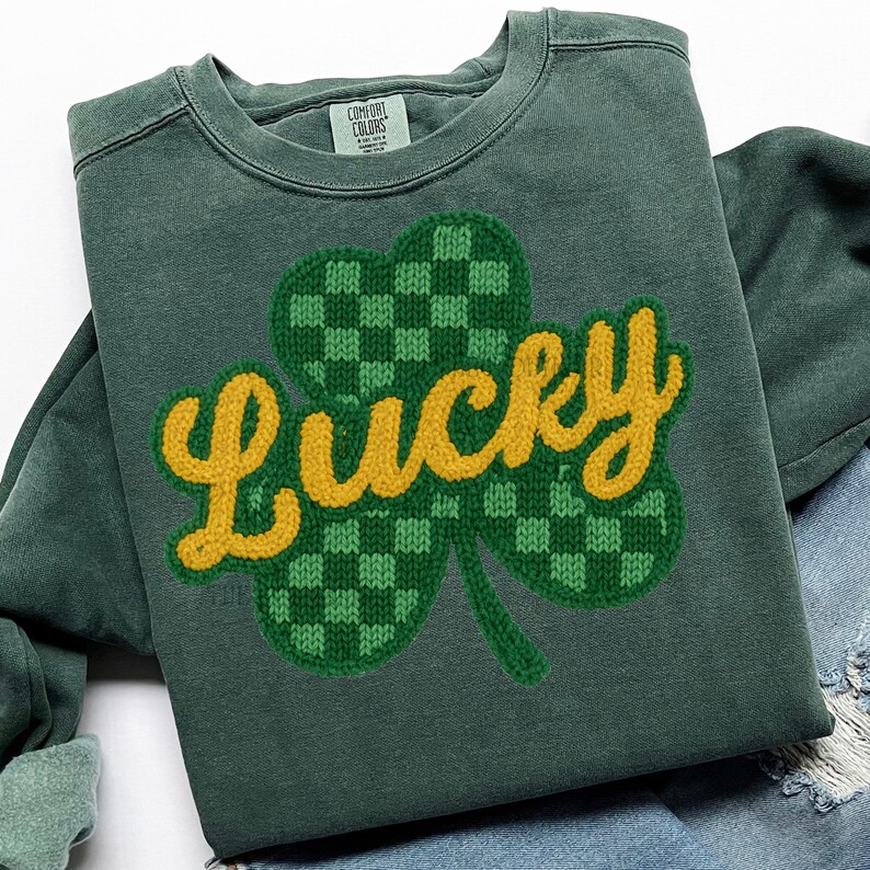 K&ouml;nnte beinhalten: Gr&uuml;nes Sweatshirt mit einem gro&szlig;en, gestickten Kleeblatt-Design. Das Kleeblatt ist gr&uuml;n mit einem Karomuster, und das Wort "Lucky" ist gelb gestickt. Das Sweatshirt ist von der Marke Comfort Colors.