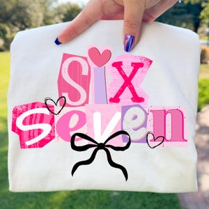 May include: A white t-shirt featuring the words "SIX SEVEN" in pink, purple, and white block letters. The design includes a black bow, hearts, and a pink heart above the letter "X".