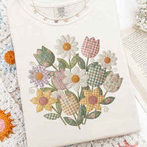 Puede incluir: Camiseta Comfort Colors color crema con un diseño de ramo floral. El ramo incluye margaritas, tulipanes y otras flores en varios patrones y colores. Un delicado collar de mariposas descansa sobre la camisa. La etiqueta dice "Comfort Colors".
