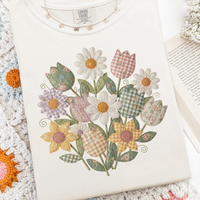 May include: Cream Comfort Colors t-shirt with a floral bouquet design. The bouquet features daisies, tulips, and other flowers in various patterns and colours. A butterfly necklace rests on the shirt. The tag reads "Comfort Colors".