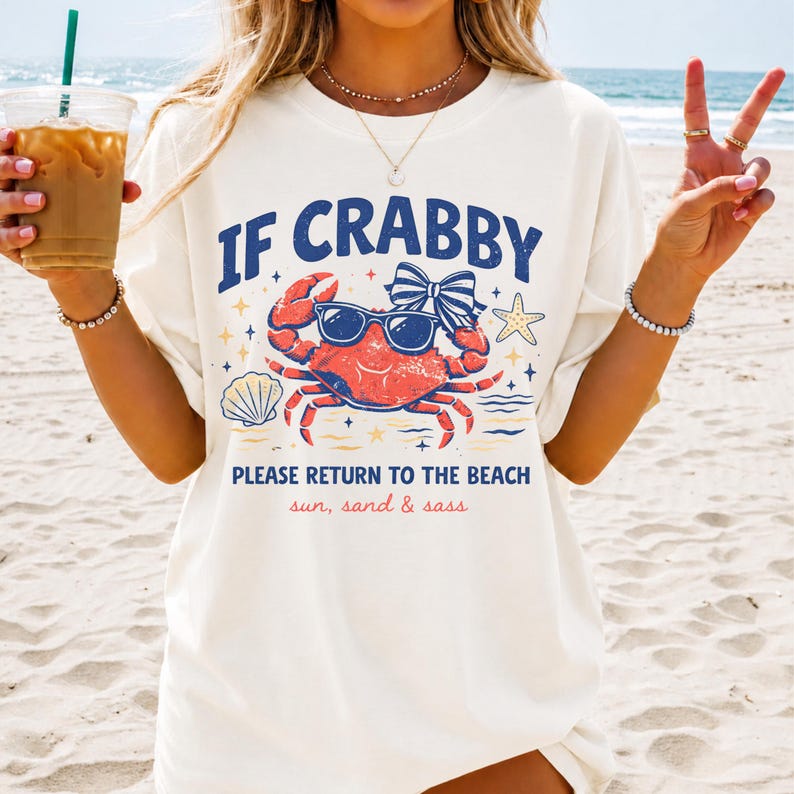 May include: Cream-colored t-shirt with the text "IF CRABBY PLEASE RETURN TO THE BEACH" and a graphic of a crab wearing sunglasses. The design includes a bow, starfish, and seashell. The words "sun, sand & sass" are also included.