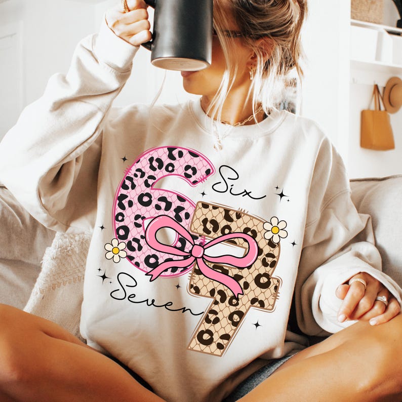 Pode incluir: Um moletom creme com um grande n&uacute;mero seis rosa com estampa de leopardo e um la&ccedil;o rosa, e um n&uacute;mero sete com estampa de leopardo. As palavras "Six" e "Seven" est&atilde;o escritas em it&aacute;lico. Pequenas flores brancas e estrelas tamb&eacute;m est&atilde;o inclu&iacute;das.