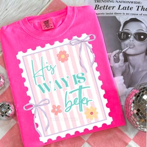 May include: Bright pink sweatshirt with a graphic that reads "His Way Is Better" in teal and white. The graphic is framed with a white border and adorned with bows and flowers. The sweatshirt is on a pink and white checkered surface.