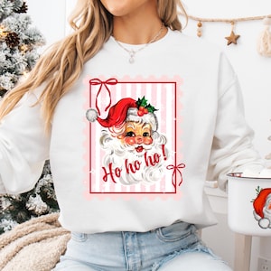 May include: White sweatshirt featuring a vintage Santa Claus graphic within a pink-striped frame. The Santa illustration has a red hat, white beard, and the words "Ho ho ho!" in red. A festive holiday-themed apparel item.