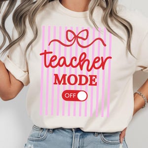 May include: Cream-colored t-shirt with a pink and white striped design. The shirt features the words "teacher MODE OFF" in red script and a red bow graphic. A fun and casual tee for teachers.