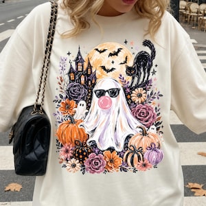 May include: A cream-colored crewneck sweatshirt featuring a graphic print of a ghost wearing black sunglasses and blowing a pink bubble. The design includes a haunted house, a black cat, bats, a full moon, pumpkins, and various purple and orange flowers.