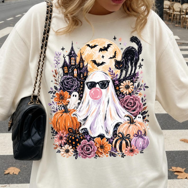 May include: A cream-colored crewneck sweatshirt featuring a graphic print of a ghost wearing black sunglasses and blowing a pink bubble. The design includes a haunted house, a black cat, bats, a full moon, pumpkins, and various purple and orange flowers.