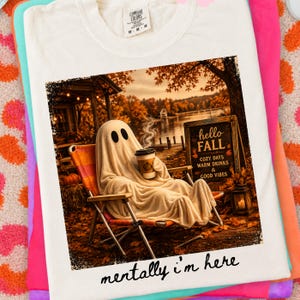 May include: Cream-colored t-shirt featuring a graphic of a ghost relaxing in a chair, holding a coffee cup, with the text "hello FALL" and "mentally i'm here". The image has a fall theme with autumn leaves and a lake in the background.