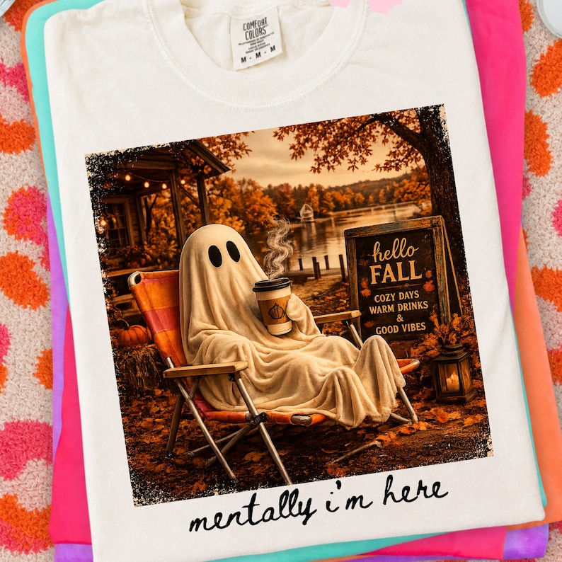 May include: Cream-colored t-shirt featuring a graphic of a ghost relaxing in a chair, holding a coffee cup, with the text "hello FALL" and "mentally i'm here". The image has a fall theme with autumn leaves and a lake in the background.
