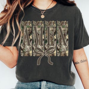 K&ouml;nnte beinhalten: Dunkelgraues T-Shirt mit dem Wort "AMEN" in einem Blockbuchstaben-Design. Die Buchstaben sind mit einem Camouflage-Muster gef&uuml;llt, und eine Schleifengrafik befindet sich unter dem Text. Das Shirt ist ein l&auml;ssiger Stil.