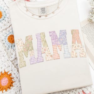 May include: Cream-colored sweatshirt with the word "MAMA" in patchwork letters. Each letter features a different floral or gingham pattern in pastel colors. A delicate butterfly necklace rests on the shirt. The brand tag "Comfort Colors" is visible.