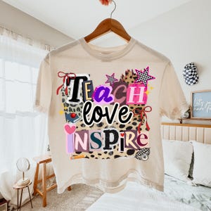May include: A beige t-shirt with the words "Teach love Inspire" in a colorful, layered design. The design includes stars, hearts, and a leopard print pattern. The shirt is hanging on a wooden hanger.