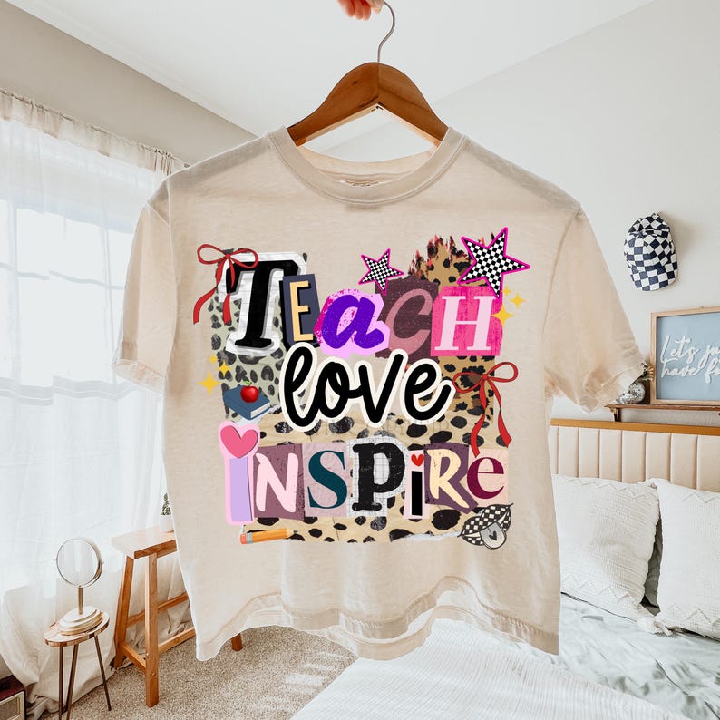 May include: A beige t-shirt with the words "Teach love Inspire" in a colorful, layered design. The design includes stars, hearts, and a leopard print pattern. The shirt is hanging on a wooden hanger.