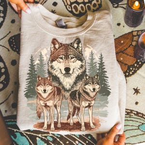 Puede incluir: Sudadera beige claro con una ilustraci&oacute;n detallada de tres lobos en un bosque. El lobo central es grande, con dos lobos m&aacute;s peque&ntilde;os a cada lado. El dise&ntilde;o incluye &aacute;rboles y una luna llena.