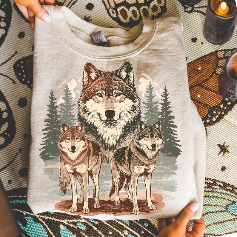 Puede incluir: Sudadera beige claro con una ilustraci&oacute;n detallada de tres lobos en un bosque. El lobo central es grande, con dos lobos m&aacute;s peque&ntilde;os a cada lado. El dise&ntilde;o incluye &aacute;rboles y una luna llena.