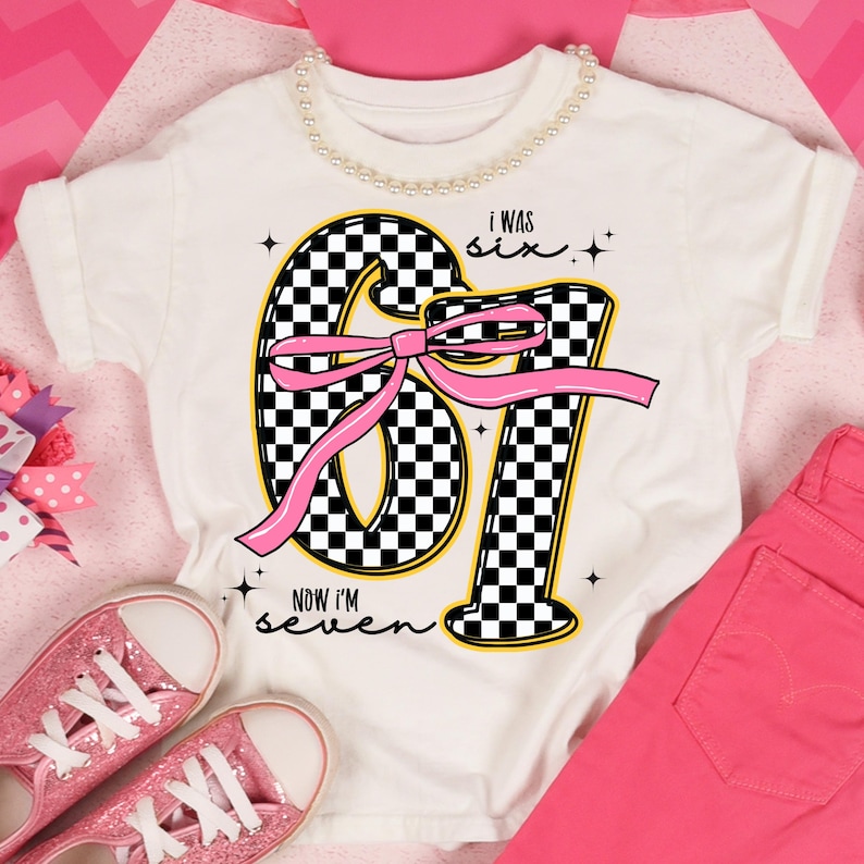 Op de afbeelding: Wit T-shirt met een grote "67" afbeelding in een zwart-wit geblokt patroon, een roze strik en de tekst "I WAS six, NOW I'M seven." Het shirt wordt gecombineerd met een roze broek en glinsterende roze sneakers.